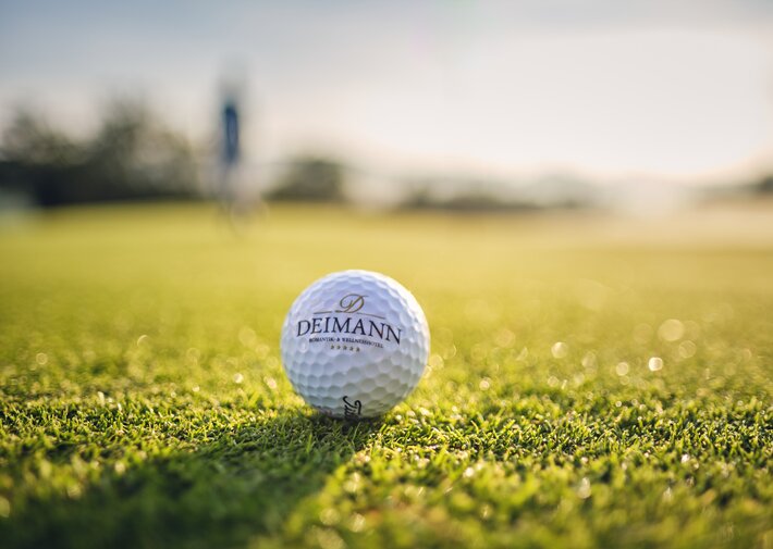 Weißer Golfball mit Deimann-Logo liegt auf grünem Golfplatzgras; Hintergrund unscharf und von warmem Sonnenlicht erhellt