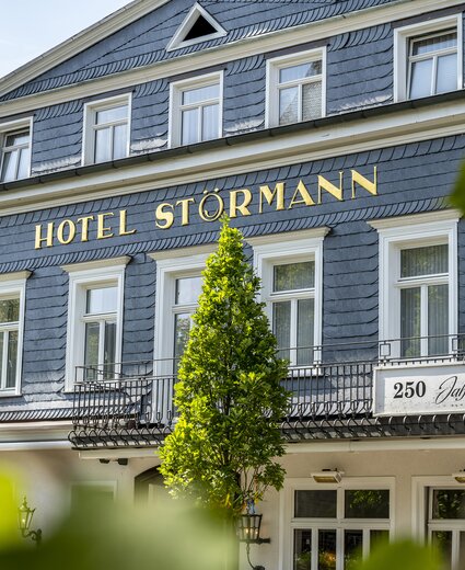 Außenansicht des Hotels Störmann mit blau-grauer Schindelfassade, goldenen Buchstaben HOTEL STÖRMANN, Balkonen und grünen Bäumen im Vordergrund.