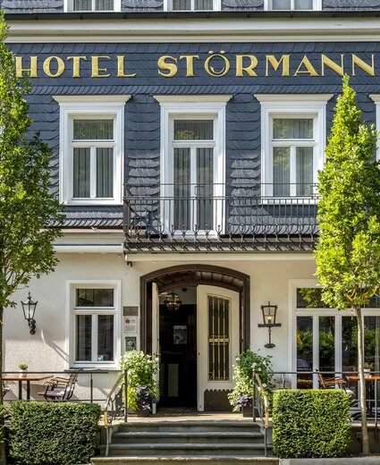 Vorderansicht des Hotel Störmann mit Eingang, Terrasse und zwei Bäumen vor der Fassade.