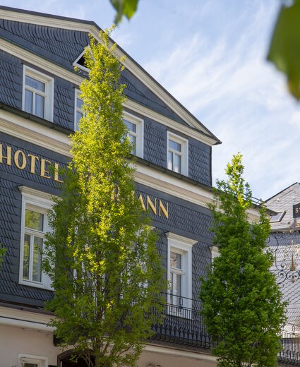 Außenansicht des Hotel Störmann mit dunkler Schieferfassade, goldener HOTEL-Schrift auf der Front, grüne Bäume davor und Schild des Restaurants am Nebengebäude.