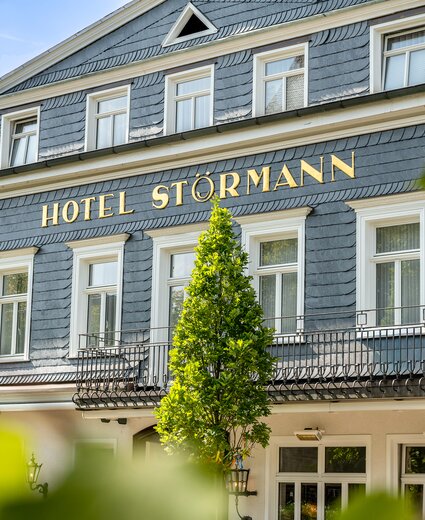 Vorderansicht des Hotels Störmann mit blau-grauem Schieferkleid, goldene Beschriftung 'HOTEL STÖRMANN', weißen Fensterrahmen, schmiedeeiserner Balkon und grünen Bäumen vor hellem Himmel.