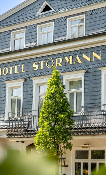 Vorderansicht des Hotels Störmann mit blau-grauem Schieferkleid, goldene Beschriftung 'HOTEL STÖRMANN', weißen Fensterrahmen, schmiedeeiserner Balkon und grünen Bäumen vor hellem Himmel.