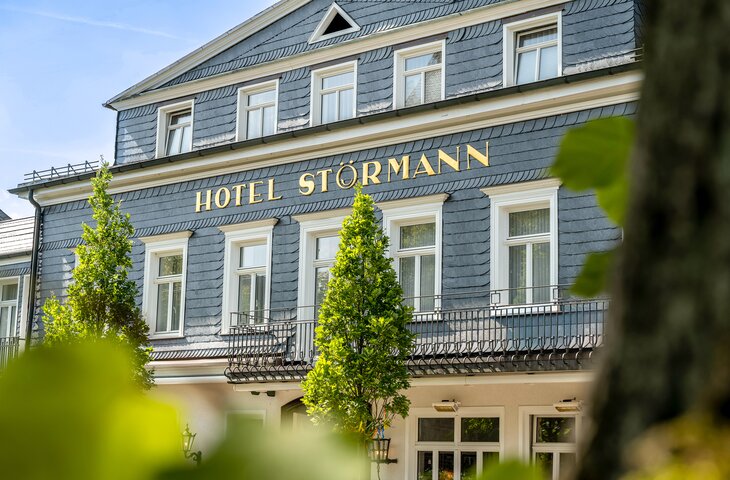 Vorderansicht des Hotels Störmann mit blau-grauem Schieferkleid, goldene Beschriftung 'HOTEL STÖRMANN', weißen Fensterrahmen, schmiedeeiserner Balkon und grünen Bäumen vor hellem Himmel.