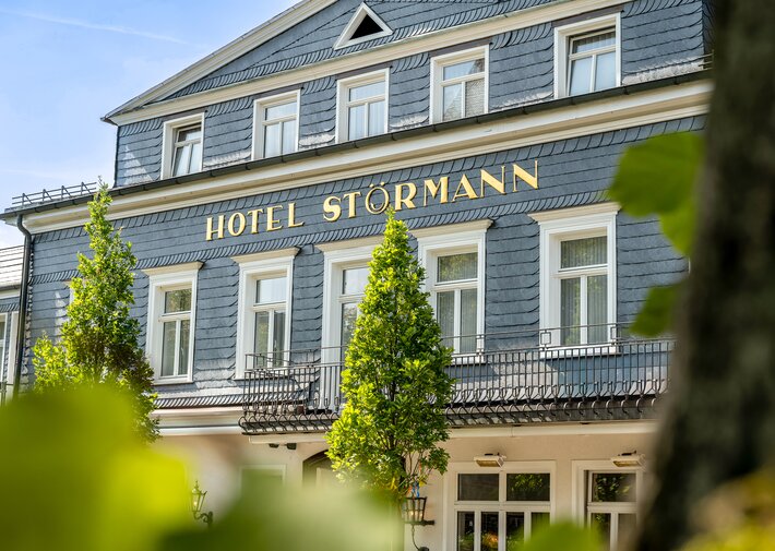 Vorderansicht des Hotels Störmann mit blau-grauem Schieferkleid, goldene Beschriftung 'HOTEL STÖRMANN', weißen Fensterrahmen, schmiedeeiserner Balkon und grünen Bäumen vor hellem Himmel.