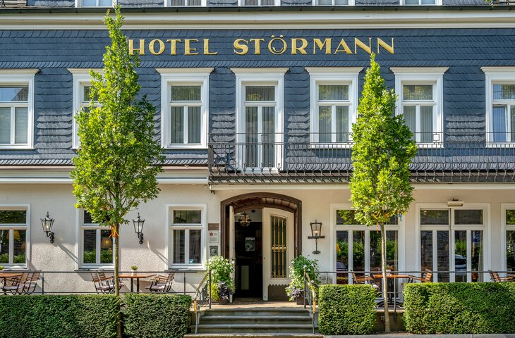 Außenansicht des Hotels Störmann: dunkles Dach, goldene Beschriftung HOTEL STÖRMANN über dem Eingang, Balkon, Eingangstür, Terrasse mit Tischen und Stühlen, zwei grüne Bäume vor der Fassade, Außenlampen und Hecken.