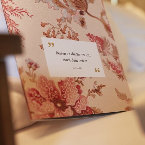 Zitatenkarte mit rosafarbigem Blumenmuster lehnt an der Bettkante; das Zitat 'Reisen ist die Sehnsucht nach dem Leben' ist lesbar; im Hintergrund ein Bettkissen.