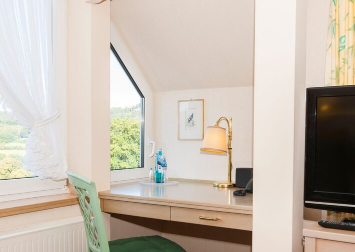 Helles Hotelzimmer mit Arbeitsbereich: Schreibtisch, Mintgrüner Stuhl, Lampe, Wasserflasche, Flachbildfernseher, Fenster mit Blick ins Grüne.