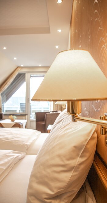 Doppelbett im Vordergrund eines Hotelzimmers; Wandlampe am Kopfteil, orange Musterwand, Sitzecke und Balkontür im Hintergrund.