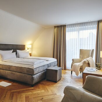 Hotelzimmer mit großem Doppelbett, Nachttischen, Leselampen, Sitzecke mit Sessel und Beistelltisch, Flachbildfernseher an der Wand, Vorhänge vor einem großen Fenster, Parkettboden.