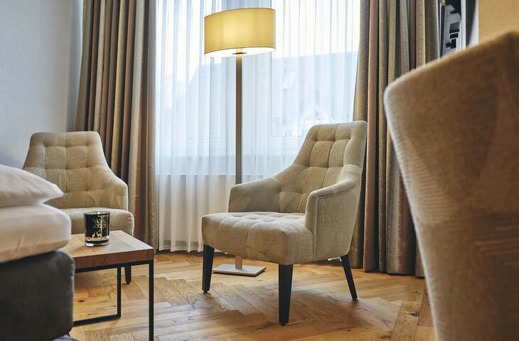 Beige gepolsterte Sessel in einer Hotel-Lounge vor Vorhängen und einem Fenster. Ein kleiner Holztisch mit einer Kerze steht davor; eine Bodenlampe sorgt für Licht.