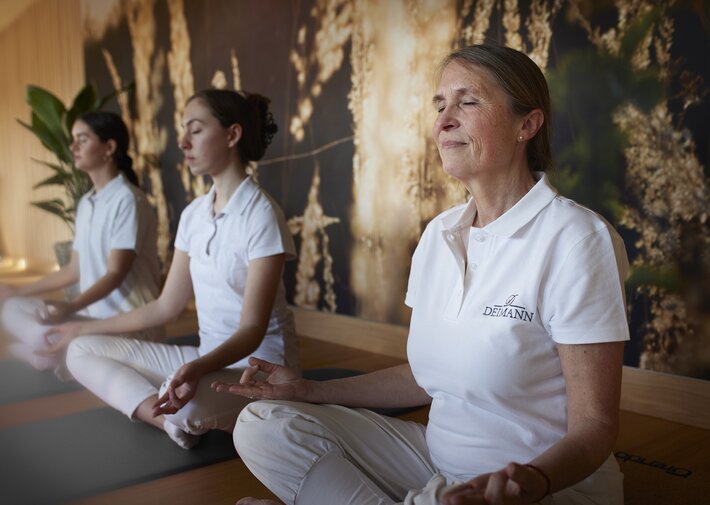Drei Frauen in weißem Yoga-Outfit sitzen auf Yogamatten im Schneidersitz in einem hellen Studio; Vordergrund eine ältere Frau mit DEIMANN-Logo auf dem Polohemd; Hintergrund zwei weitere meditierende Frauen; Naturwand, Pflanzen und Holzboden.