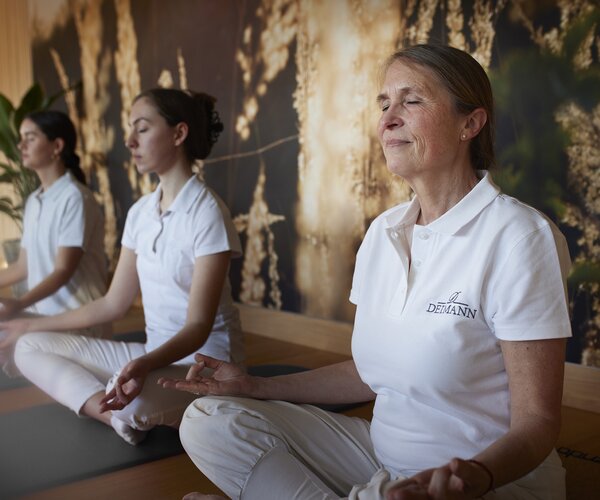 Drei Frauen in weißem Yoga-Outfit sitzen auf Yogamatten im Schneidersitz in einem hellen Studio; Vordergrund eine ältere Frau mit DEIMANN-Logo auf dem Polohemd; Hintergrund zwei weitere meditierende Frauen; Naturwand, Pflanzen und Holzboden.