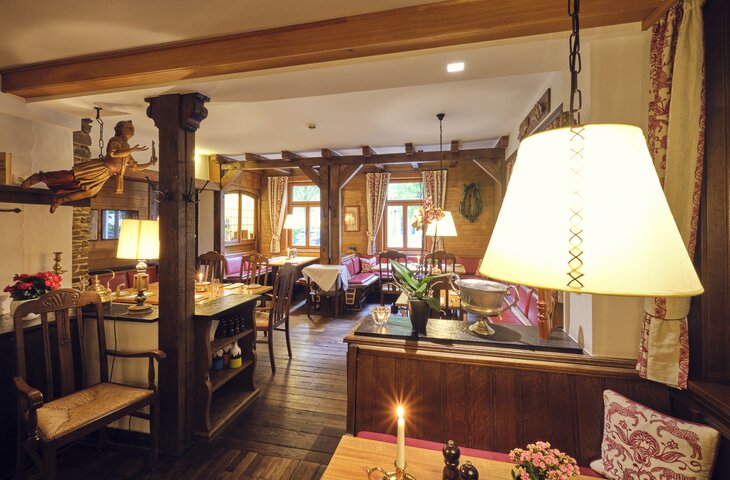 Rustikaler Innenraum eines Eventlokals mit dunklen Holzbalken, Holzfußboden, roter gepolsterter Sitzbereich, Tische mit Kerzen und Blumen, Lampen und warme Beleuchtung, Fenster mit Vorhängen.