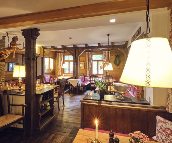 Rustikaler Innenraum eines Eventlokals mit dunklen Holzbalken, Holzfußboden, roter gepolsterter Sitzbereich, Tische mit Kerzen und Blumen, Lampen und warme Beleuchtung, Fenster mit Vorhängen.