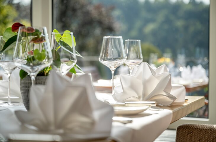 Gedeckter Tisch im Restaurant des Hotel Störmann mit gefalteter weißer Serviette, Weingläsern und Pflanzenschmuck, helles Tageslicht, Blick nach draußen.