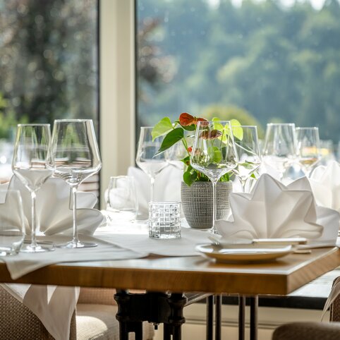 Gedeckter Esstisch im Hotel Störmann im Sauerland: gefaltete weiße Servietten, Wein- und Wassergläser, Pflanze in einer Vase, vor einem großen Fenster mit Blick ins Grüne.
