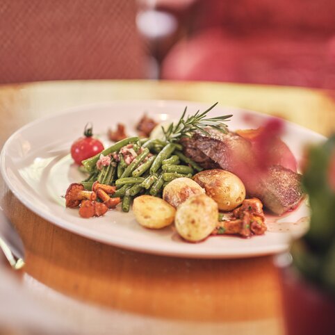 Weißer Teller mit grünen Bohnen, Kartoffeln, Speckwürfeln, einer Fleischscheibe, Kirschtomate und Rosmarin, serviert auf einem Holztisch