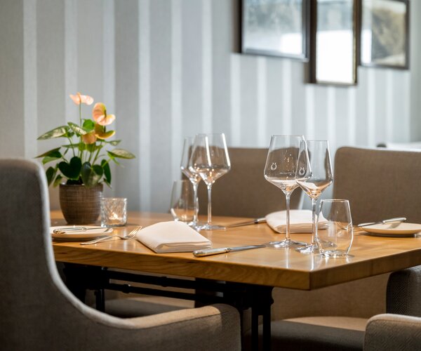 Gedeckter Holztisch in einem modernen Hotelrestaurant mit zwei Weingläsern, einem Wasserglas, Tellern, Besteck, Stoffservietten und einer Pflanzendekoration; graue gepolsterte Stühle und vertikale Wandverkleidung im Hintergrund.