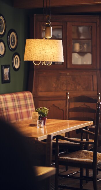 Innenaufnahme eines rustikalen Hotel-Esszimmers: Holztisch mit Stühlen, eine plaid-gepolsterte Bank links, eine hängende Leuchte mit Stoffschirm; Hintergrund ein großer Holzschrank mit Glastüren; Tischplatte mit Pflanze, Kerze und Tischkarte; grüne Wand mit runden Bilderrahmen.