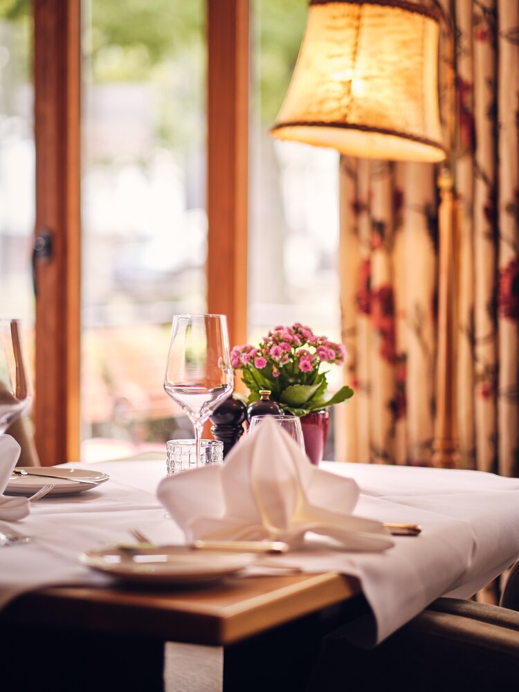 Gedeckter Restauranttisch mit weißem Tischtuch, gefalteter Serviette, Weinglas, Wasserglas und einer kleinen Topfpflanze mit pinkfarbenen Blüten; im Hintergrund Holzfensterrahmen, florale Vorhänge und warmes Lampenlicht.