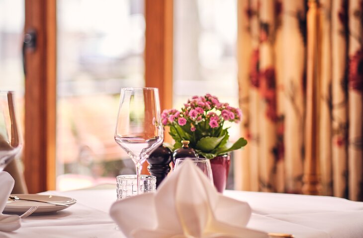 Gedeckter Restauranttisch mit weißem Tischtuch, gefalteter Serviette, Weinglas, Wasserglas und einer kleinen Topfpflanze mit pinkfarbenen Blüten; im Hintergrund Holzfensterrahmen, florale Vorhänge und warmes Lampenlicht.