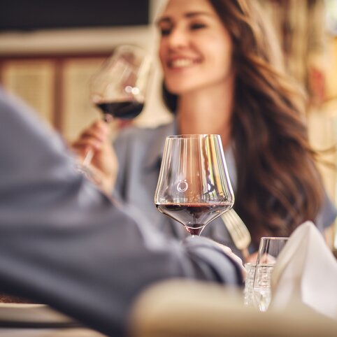 Frau mit langen braunen Haaren lächelt und hält ein Glas Rotwein; im Vordergrund ein großes Weinglas mit Rotwein; am Tisch liegen Gläser, Besteck und eine gefaltete Serviette.