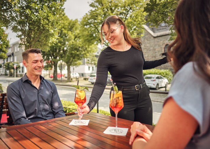 Eine Kellnerin serviert einem Mann im Straßencafé einen orangefarbenen Cocktail; eine weitere Person von hinten im Vordergrund; zwei Cocktails stehen auf dem Holztisch, Hintergrund mit Bäumen und Gebäuden.