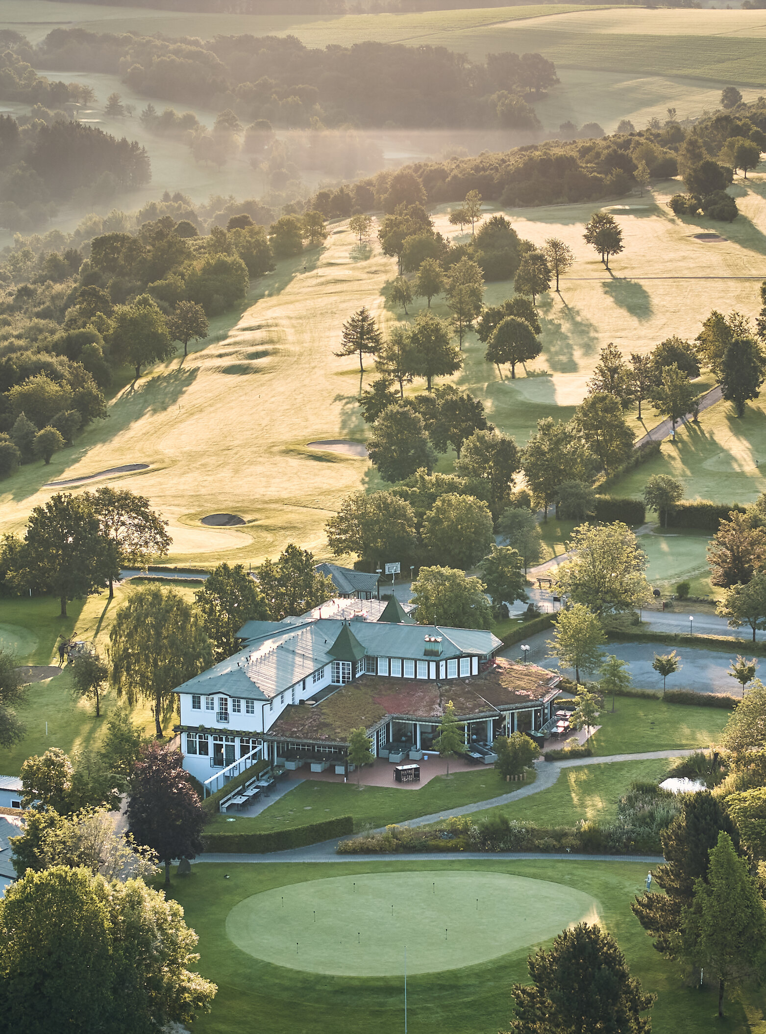 Vogelperspektive auf einen Golfplatz mit Clubhaus im Vordergrund, grünen Grüns, vielen Bäumen, Sandbunkern und Wegen; sanftes Morgenlicht und weiter Landschaft im Hintergrund.