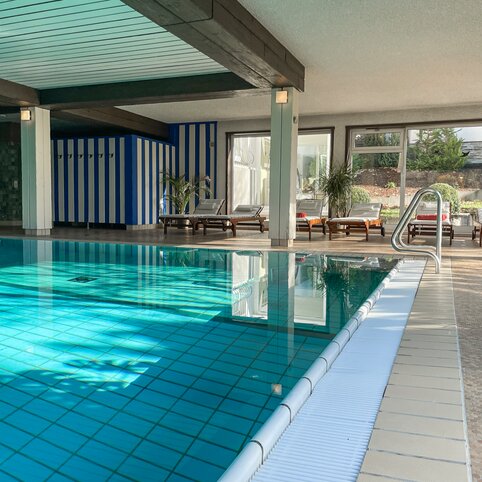 Innenpoolbereich eines Hotels mit türkisfarbenem Wasser, rechteckiges Becken, Fliesenboden, Liegen mit weißen Auflagen an der Fensterfront, Pflanzen, Edelstahl-Handlauf am Beckenrand, blau-weiß gestreifter Wandabschnitt, dunkle Holzbalken an der Decke.