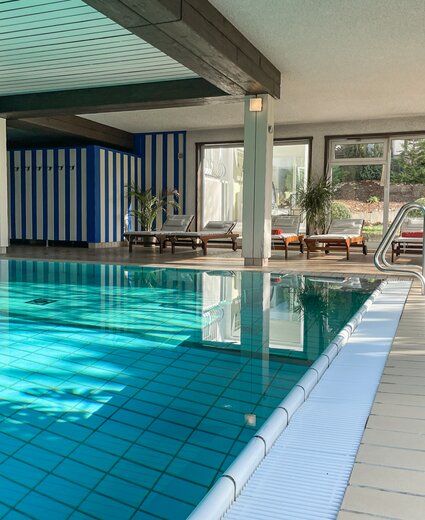 Innenpoolbereich eines Hotels mit türkisfarbenem Wasser, rechteckiges Becken, Fliesenboden, Liegen mit weißen Auflagen an der Fensterfront, Pflanzen, Edelstahl-Handlauf am Beckenrand, blau-weiß gestreifter Wandabschnitt, dunkle Holzbalken an der Decke.