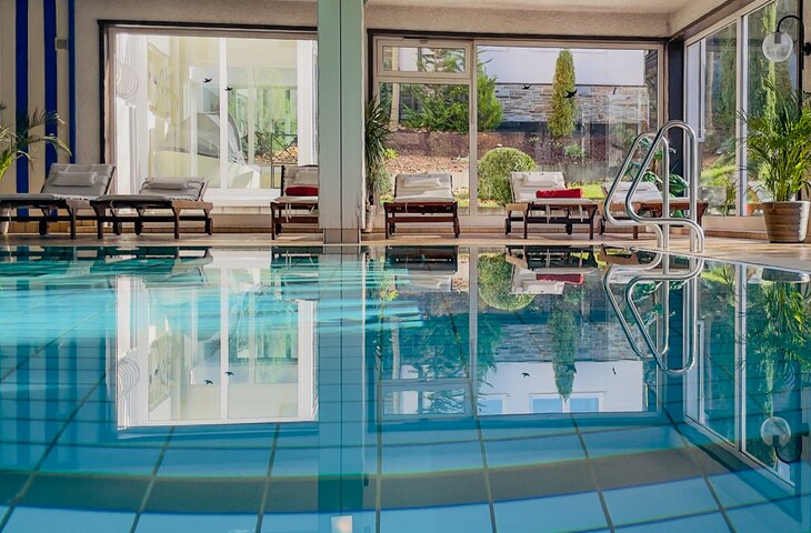 Innenpoolbereich eines Hotels mit türkisfarbenem Becken, weißen Liegenauflagen, großen Glasfenstern und Topfpflanzen; Reflexionen der Umgebung auf der Wasseroberfläche.