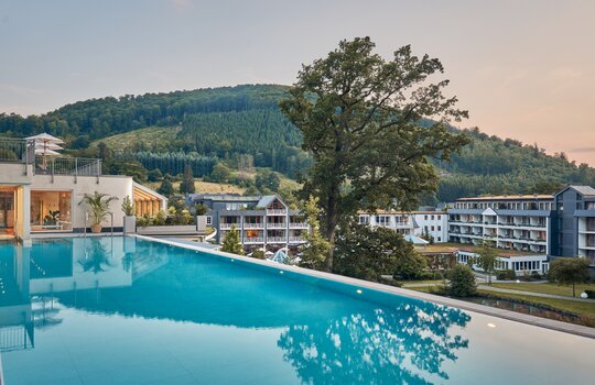 Außenansicht eines modernen Hotels mit Infinity-Pool im Vordergrund; Hintergrund grüne Hügel und Wälder, Baum steht in der Mitte, weitere Hotelgebäude dahinter.