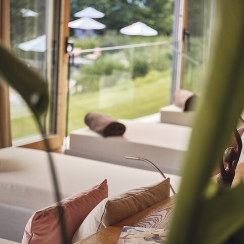 Wellness-Lounge im Hotel Störmann: Zwei weiße Liegen mit braunen Handtüchern und Kissen, ein Holztisch mit Dekoration, Blick durch große Fenster auf eine grüne Gartenlandschaft, Vorhänge und eine Pflanze im Vordergrund.