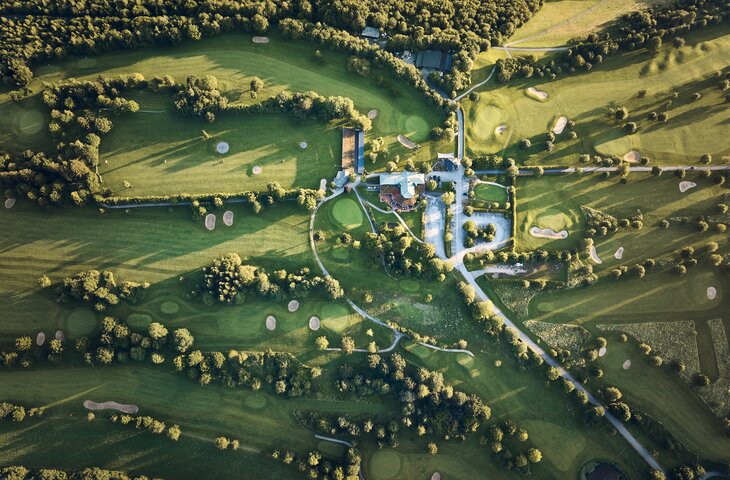 Vogelperspektive eines Golfplatzes mit zentralem Gebäudekomplex, von Bäumen umgeben, Sandbunkern und Wegen.