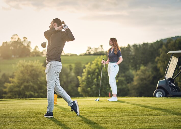 Mann beim Golfschwung auf grünem Golfplatz; Frau beobachtet mit Golfschläger; Golfwagen rechts; Bäume und Abendlicht im Hintergrund.
