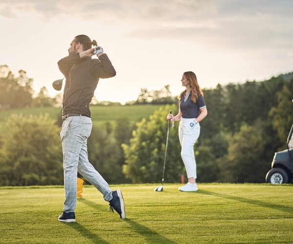 Mann beim Golfschwung auf grünem Golfplatz; Frau beobachtet mit Golfschläger; Golfwagen rechts; Bäume und Abendlicht im Hintergrund.
