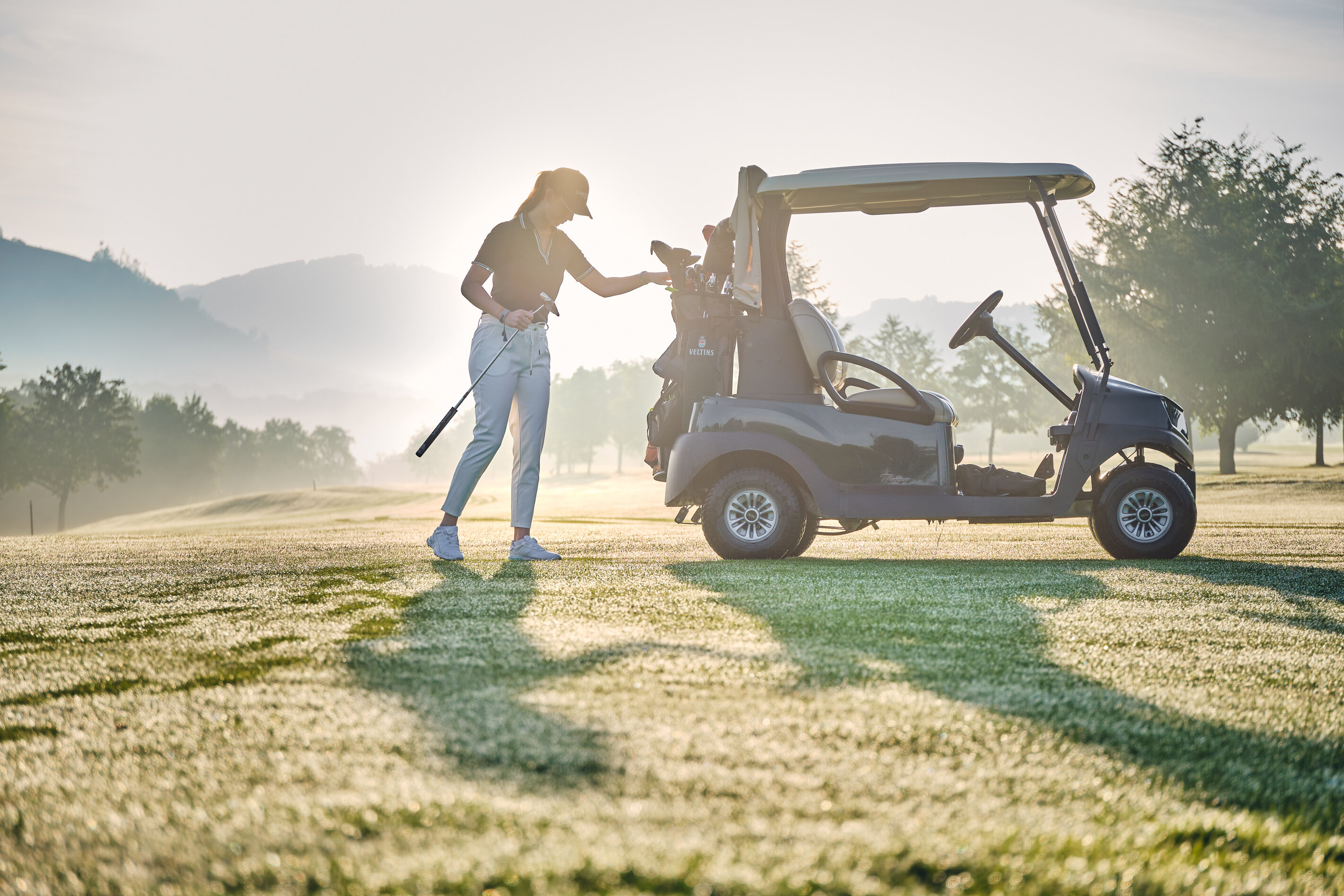 Eine Person im Golfoutfit steht neben einem geparkten Golfwagen auf einem Golfplatz; im Hintergrund sind Bäume und sanfte Hügel bei Morgensonne zu sehen.