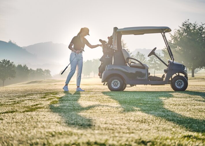 Eine Person im Golfoutfit steht neben einem geparkten Golfwagen auf einem Golfplatz; im Hintergrund sind Bäume und sanfte Hügel bei Morgensonne zu sehen.