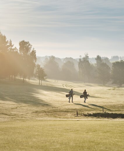 Zwei Golfer mit Golftaschen gehen über einen offenen Golfplatz; im Hintergrund Bäume, Sonnenstrahlen durch Nebel, weite Grasflächen