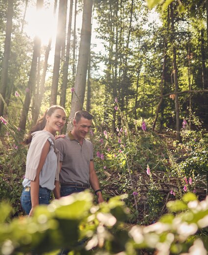 Paar im Wald: Eine Frau und ein Mann gehen nebeneinander; Sonnenlicht fällt durch Bäume; violette Blüten am Weg; unscharfer Vordergrund.