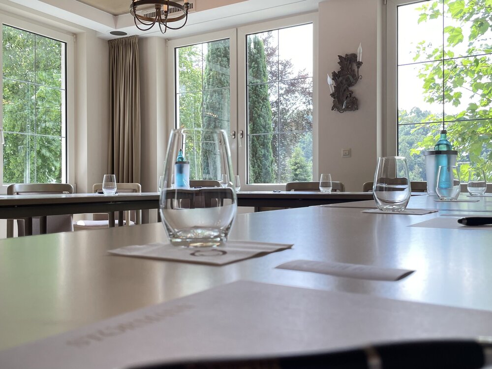 Konferenzraum mit großen Fenstern und Blick ins Grüne; Tische in U-Form mit Wasserflaschen, Gläsern, Notizblättern und Stiften; beige Vorhänge und Deckenleuchte