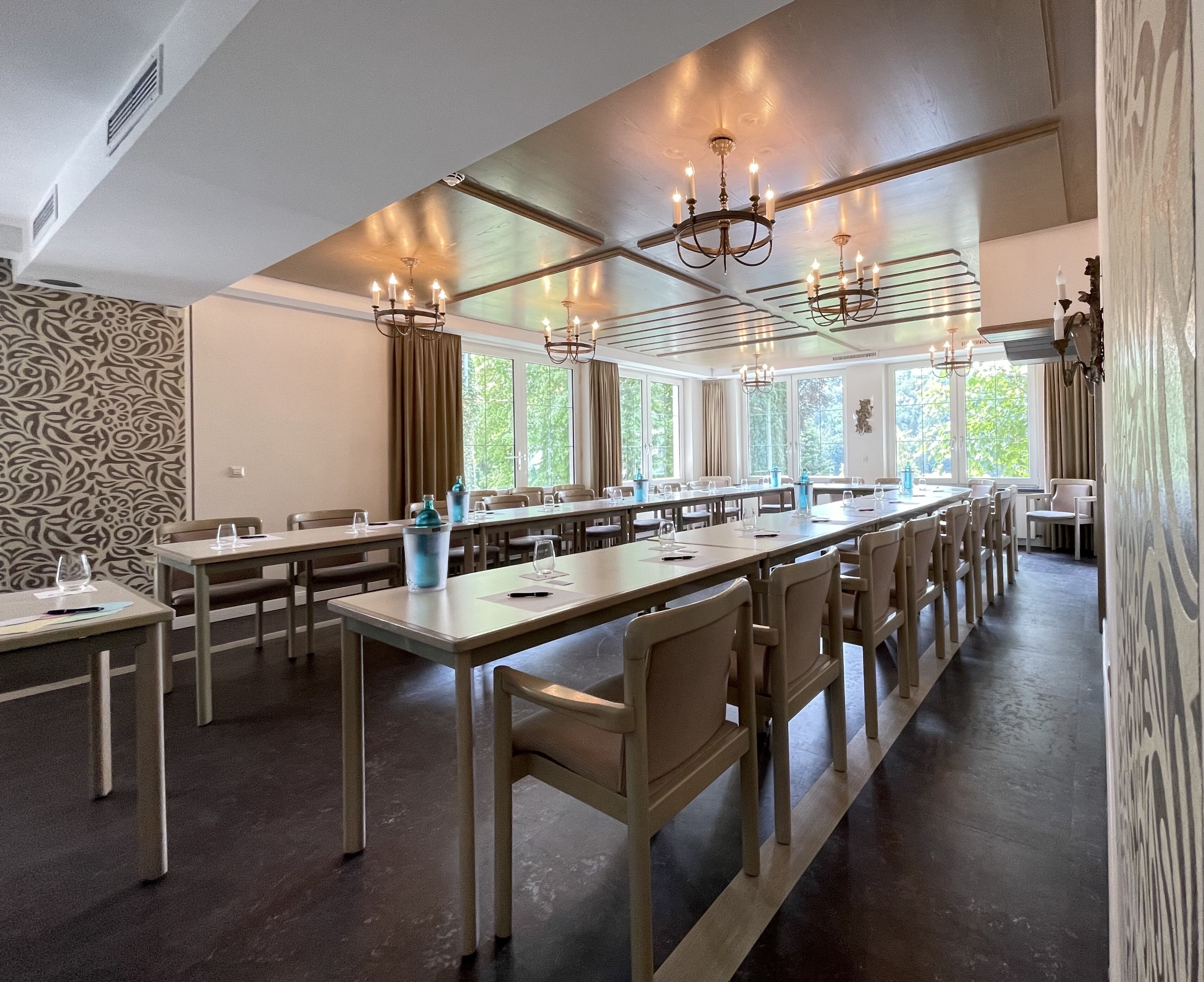 Konferenzraum mit parallelen Tischen und beige Stühlen, Kronleuchter an der Decke, große Fensterfront mit Vorhängen, Wasserflaschen und Gläser auf den Tischen, gemusterte Wände.