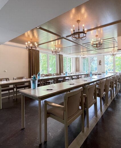 Konferenzraum mit parallelen Tischen und beige Stühlen, Kronleuchter an der Decke, große Fensterfront mit Vorhängen, Wasserflaschen und Gläser auf den Tischen, gemusterte Wände.